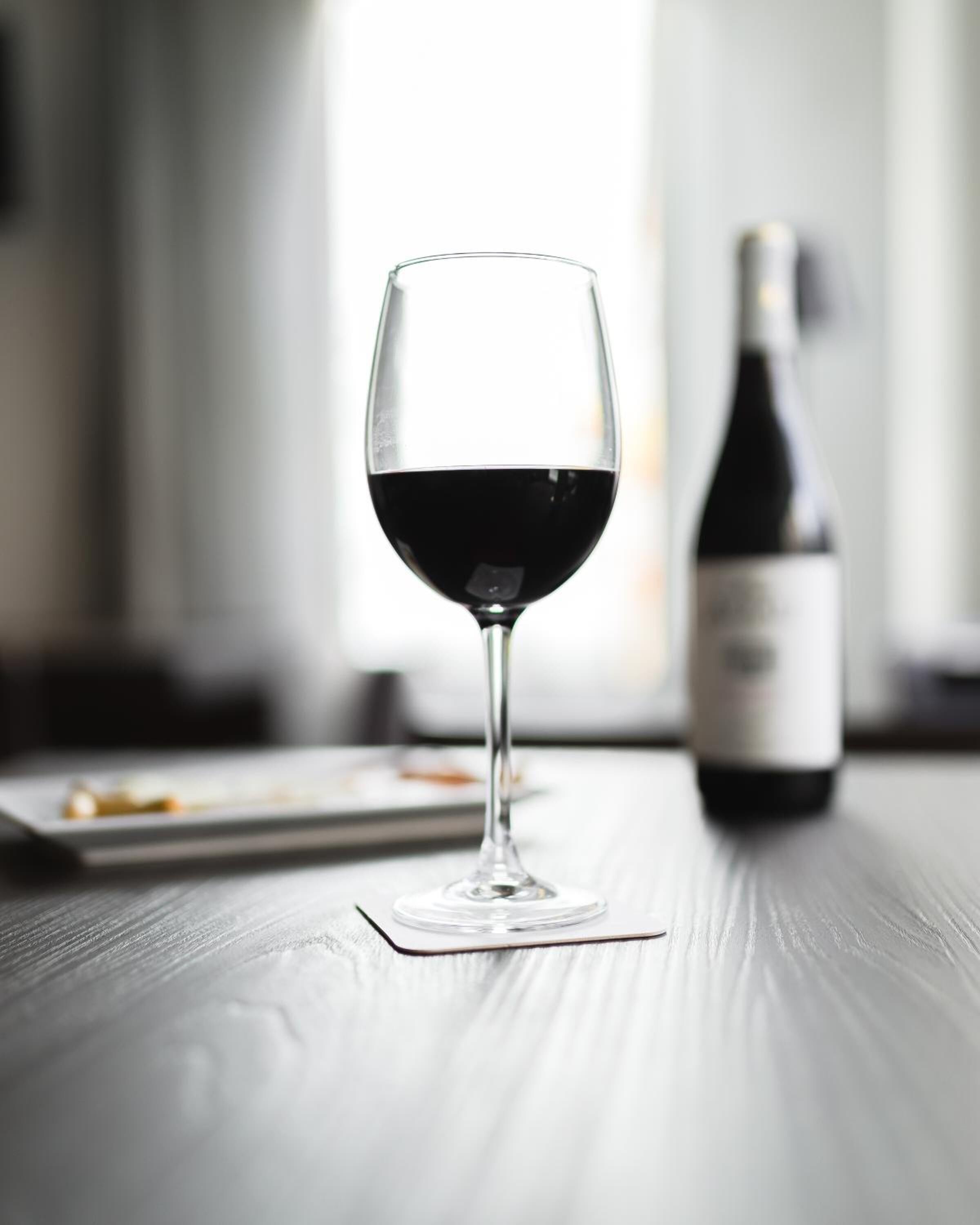 A wine glass filled with red wine on a wooden table with a bottle in the background.