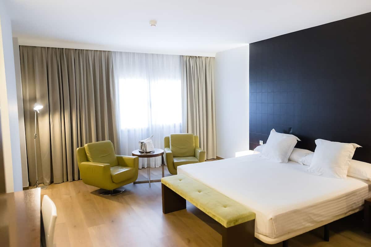 Une chambre d’hôtel avec un grand lit, deux fauteuils, une petite table et une lampe.