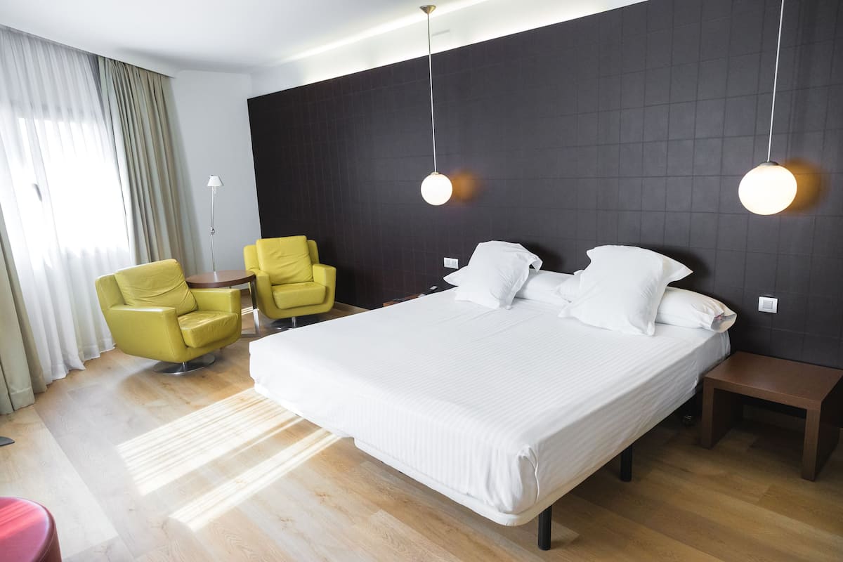 Une chambre d’hôtel moderne avec un grand lit, deux tables de chevet, un bureau et deux fauteuils jaunes.