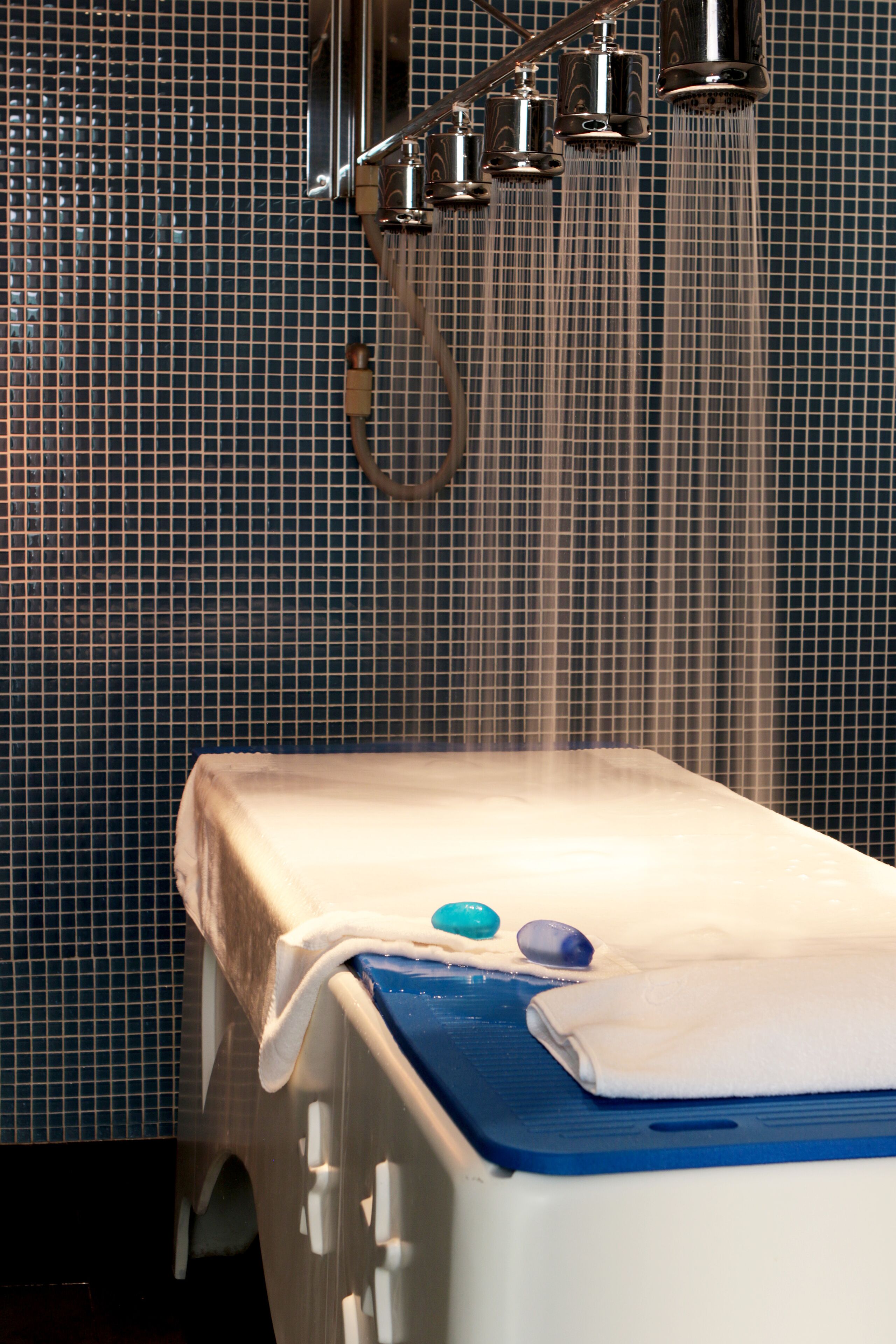 Una bañera con un tapete azul y una toalla blanca, un cabezal de ducha encima, y una pared de azulejos al fondo.