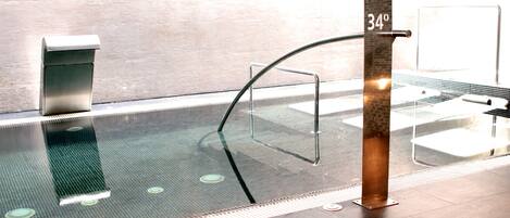 Piscina interior moderna com paredes de vidro transparente, guarda-corpo de metal e indicador de temperatura de 34 graus.