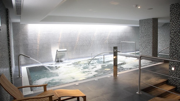Un área de spa moderna con una bañera de hidromasaje, asientos de madera y una pared de azulejos.