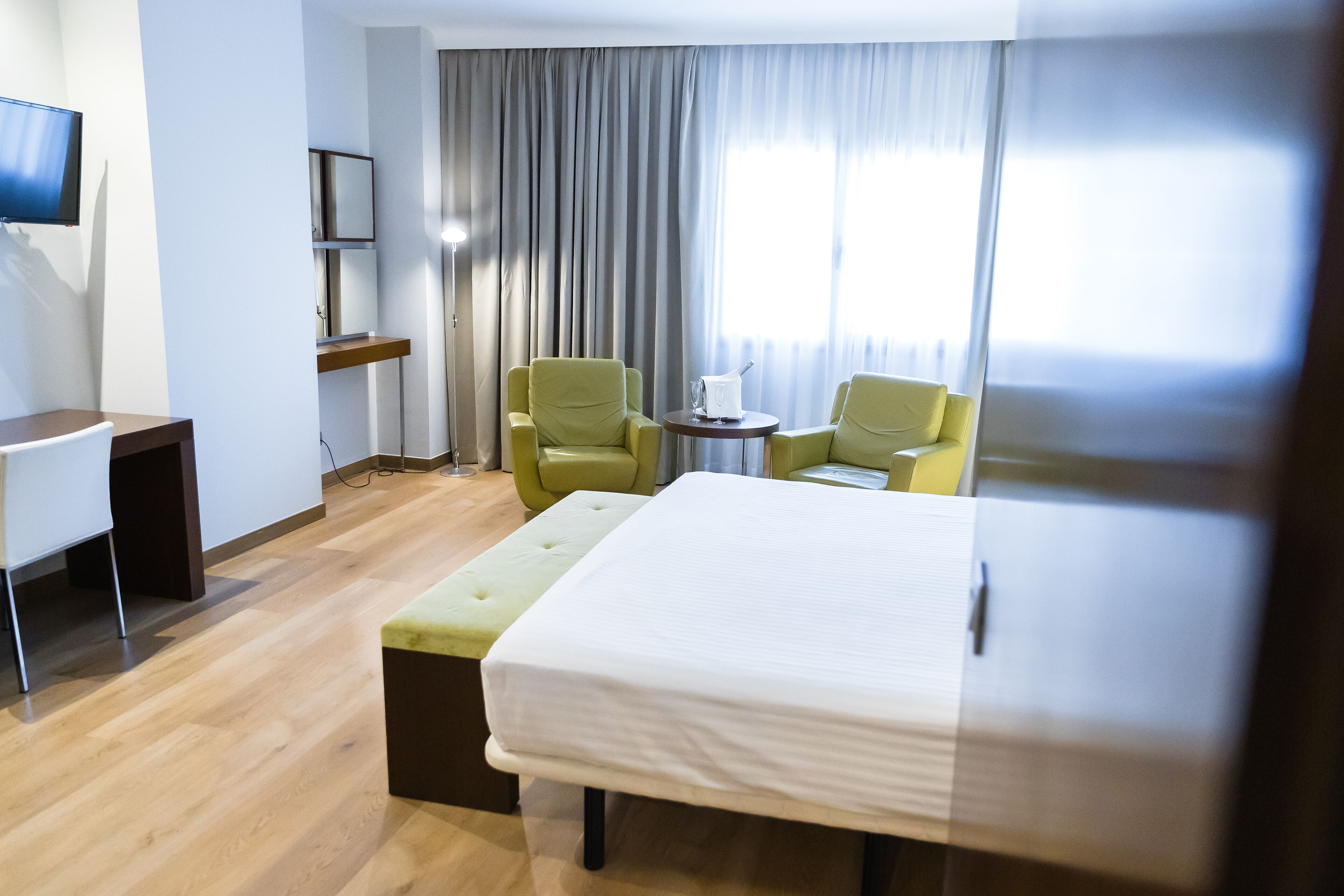 A hotel room with a bed, two armchairs, a small table, a desk, and a TV.