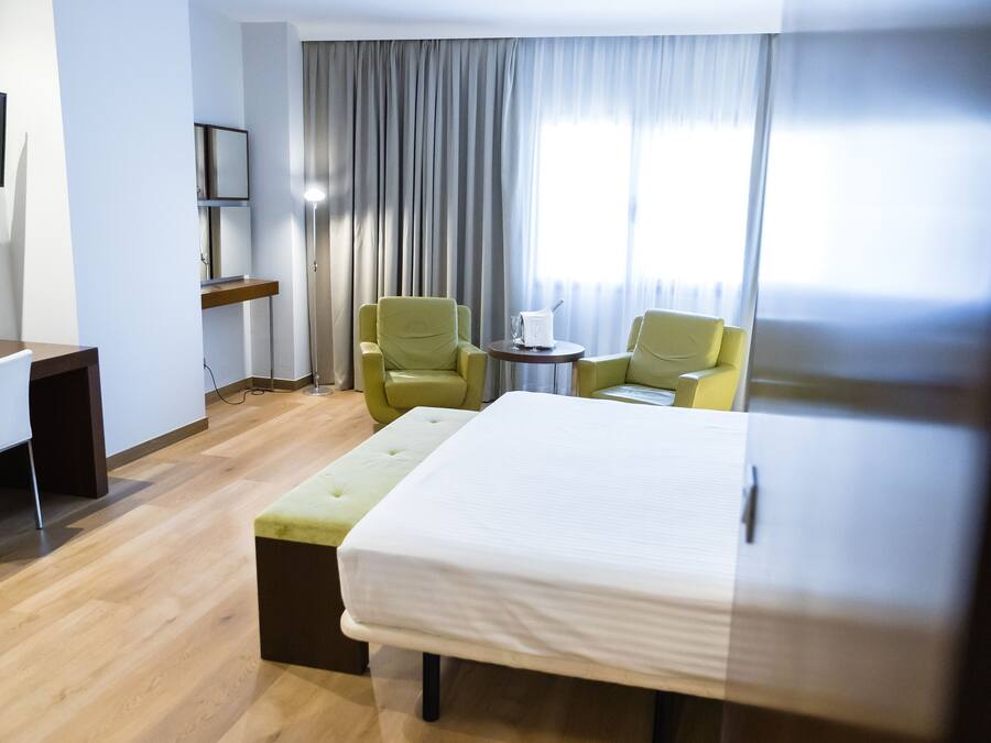 A hotel room with a bed, two armchairs, a small table, a desk, and a TV.
