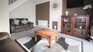 Una sala de estar con un sofá de cuero marrón, una mesa de centro de madera, un mueble bar con puertas de cristal y un televisor montado en la pared.