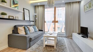 A modern living room with a grey sofa, white coffee table, and a TV on a white cabinet. There are framed pictures on the shelves and a large window with curtains. - Flats For Rent - Aura Spa&Wellness (Gdansk)