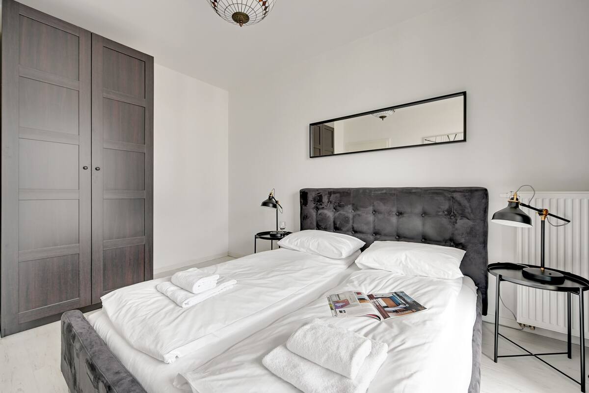 Ein ordentlich bezogenes Bett mit weißen Leinen und Handtüchern, ein dunkles Kopfteil, ein Nachttisch mit einer Lampe und ein großer Spiegel an der Wand.