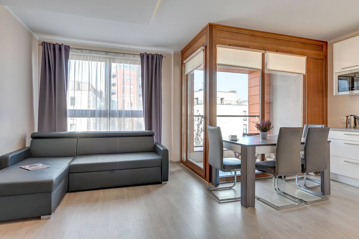 Ein modernes Wohnzimmer mit einer grauen Eckcouch, einem Esstisch mit Stühlen und einer Schiebeglastür, die auf einen Balkon führt.