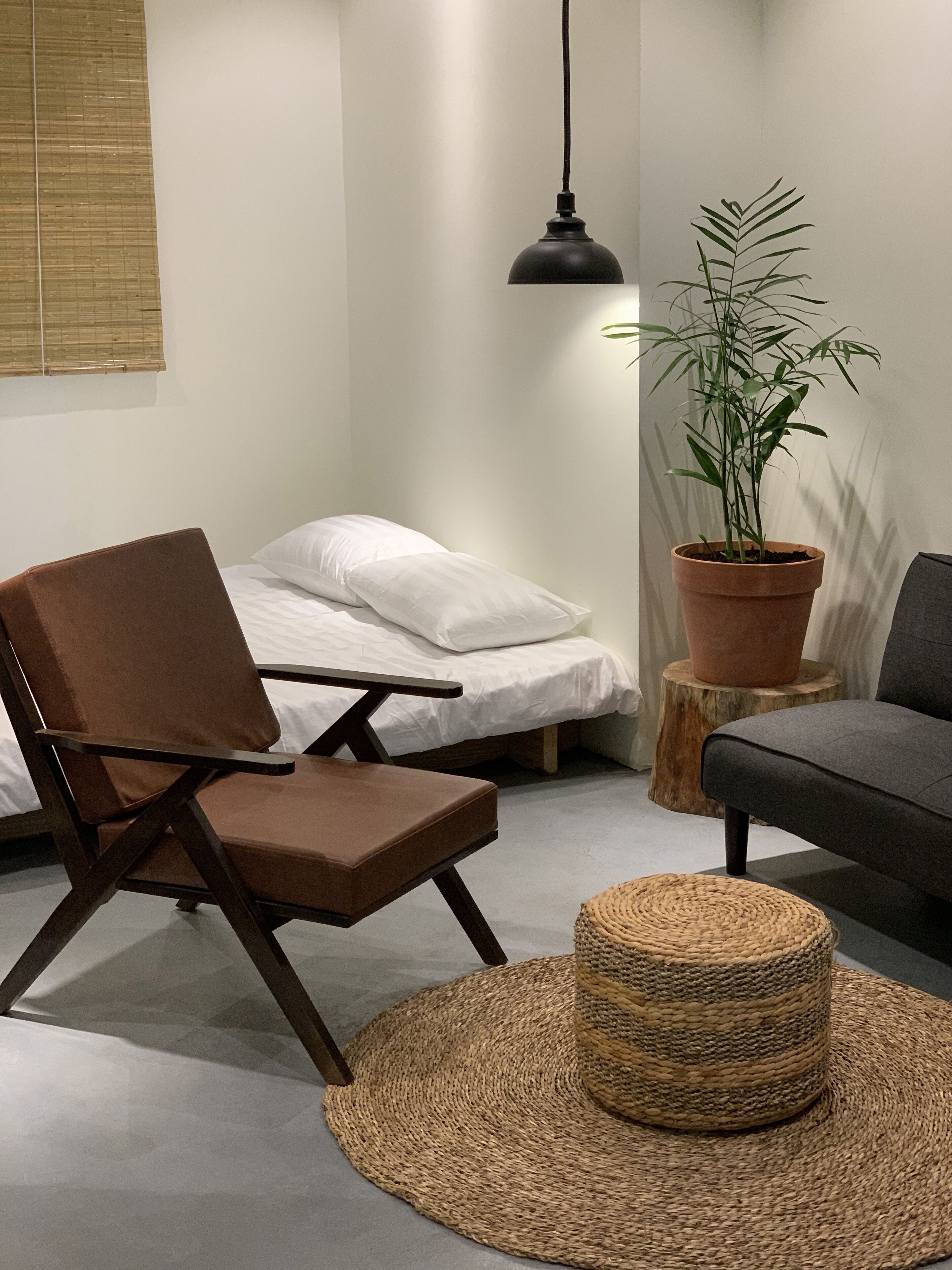 A room with a bed, a brown chair, a grey sofa, a woven ottoman, a potted plant, and a hanging lamp.