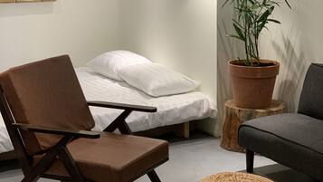 A room with a bed, a brown chair, a grey sofa, a woven ottoman, a potted plant, and a hanging lamp.