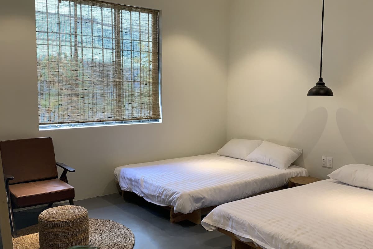 A modern bedroom with two single beds, a wooden chair, a small round table, and a large window with blinds.