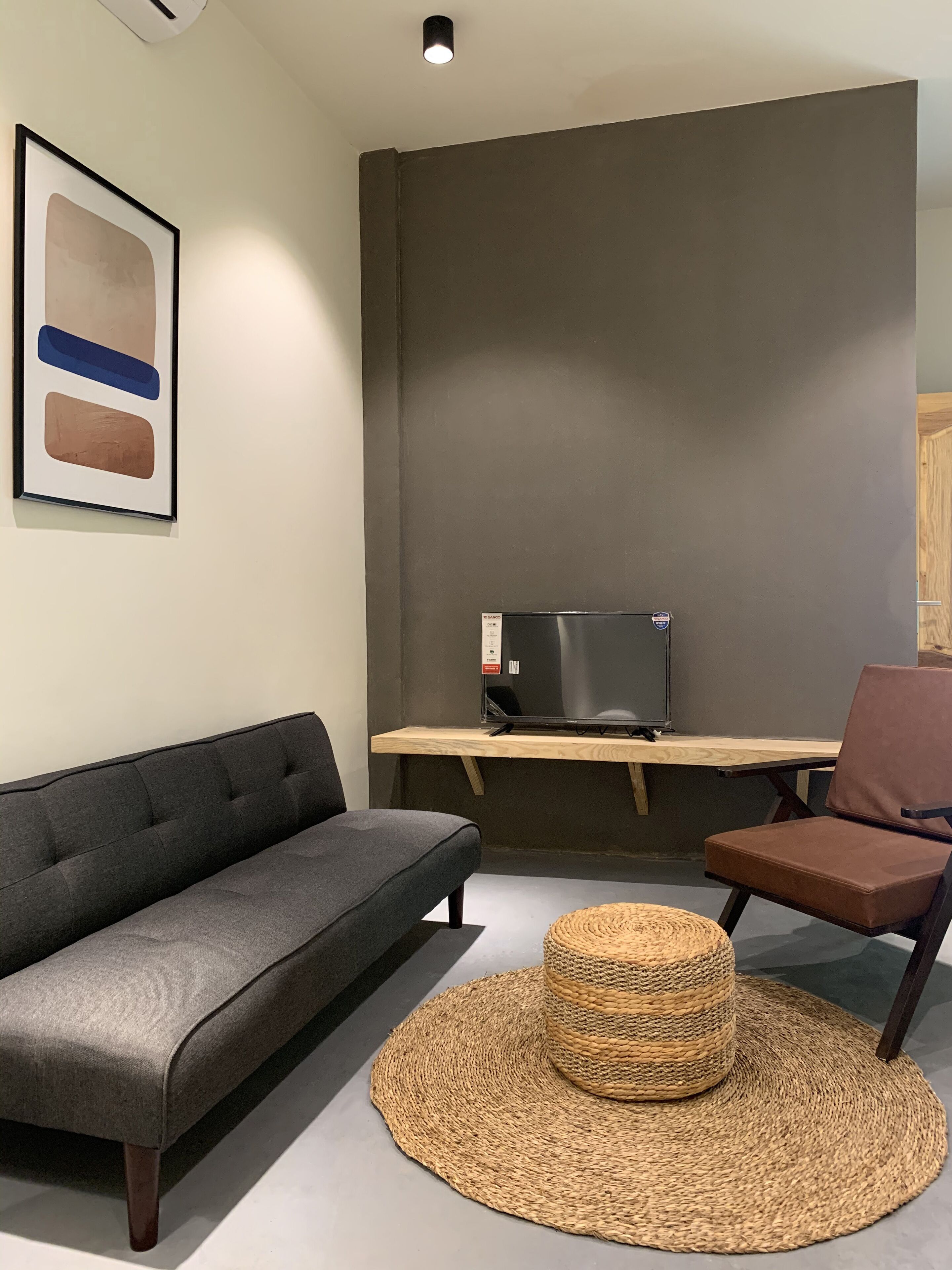 A modern living room with a grey sofa, a brown chair, a woven ottoman, a wooden desk with a flat-screen TV, and a framed abstract artwork on the wall.