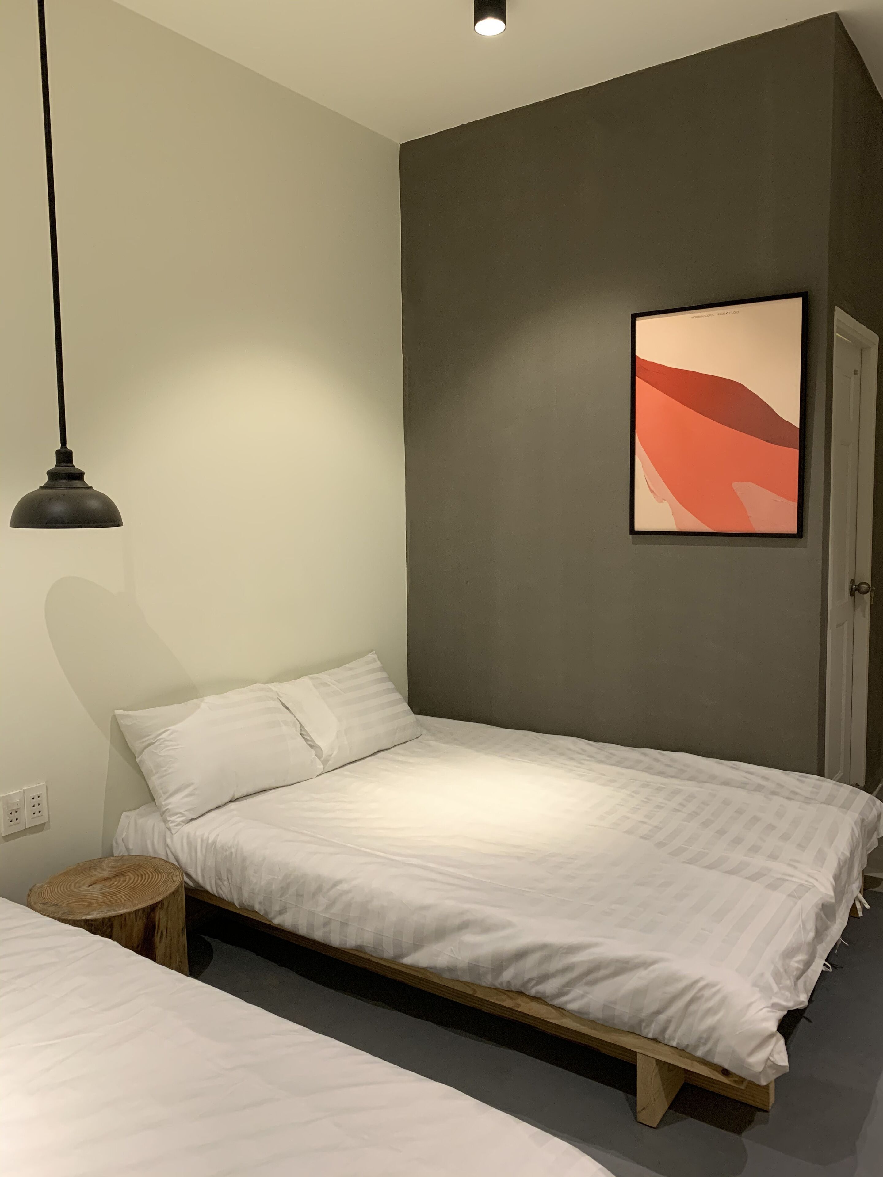 A bedroom with a single bed, a small wooden stool, a wall-mounted light, and a framed abstract painting.