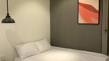 A bedroom with a single bed, a small wooden stool, a wall-mounted light, and a framed abstract painting.
