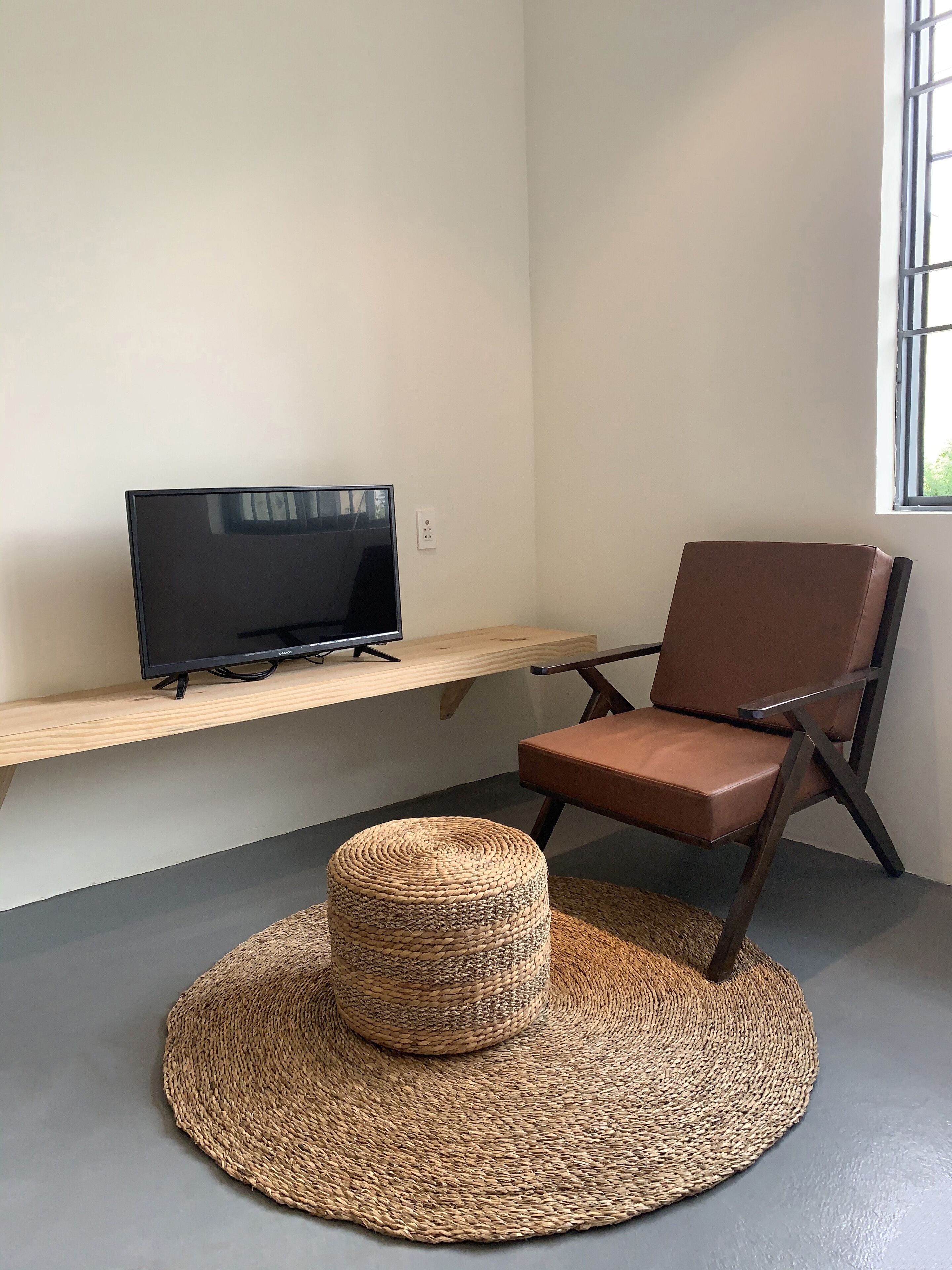A room with a TV on a wooden shelf, a brown leather chair, and a woven ottoman on a grey floor.