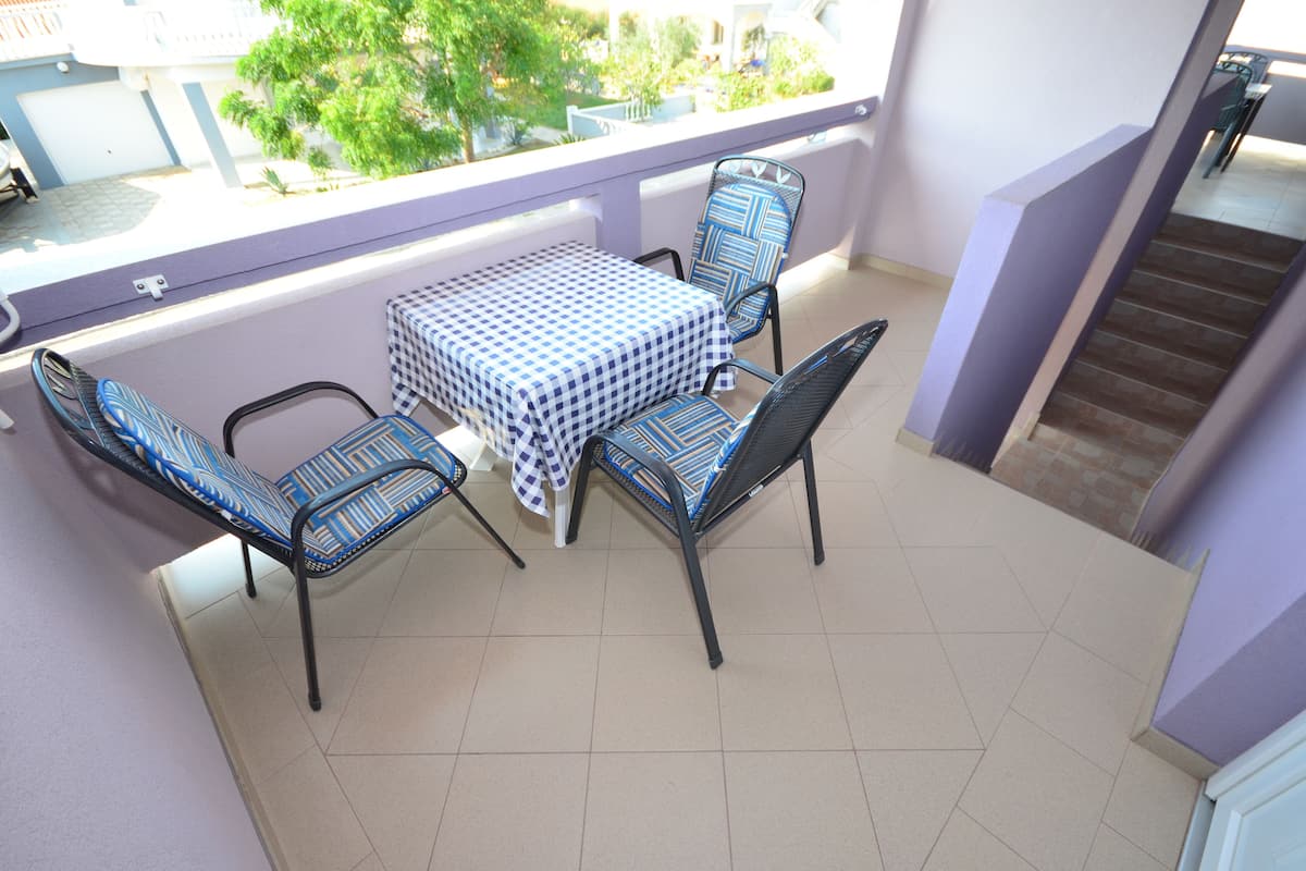 A balcony with a checkered table and chairs, a tiled floor, and a view of greenery and buildings.