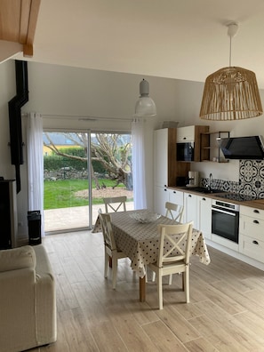 Uma cozinha moderna com armários brancos, uma mesa de jantar com toalha xadrez e uma janela grande com vista para um jardim.