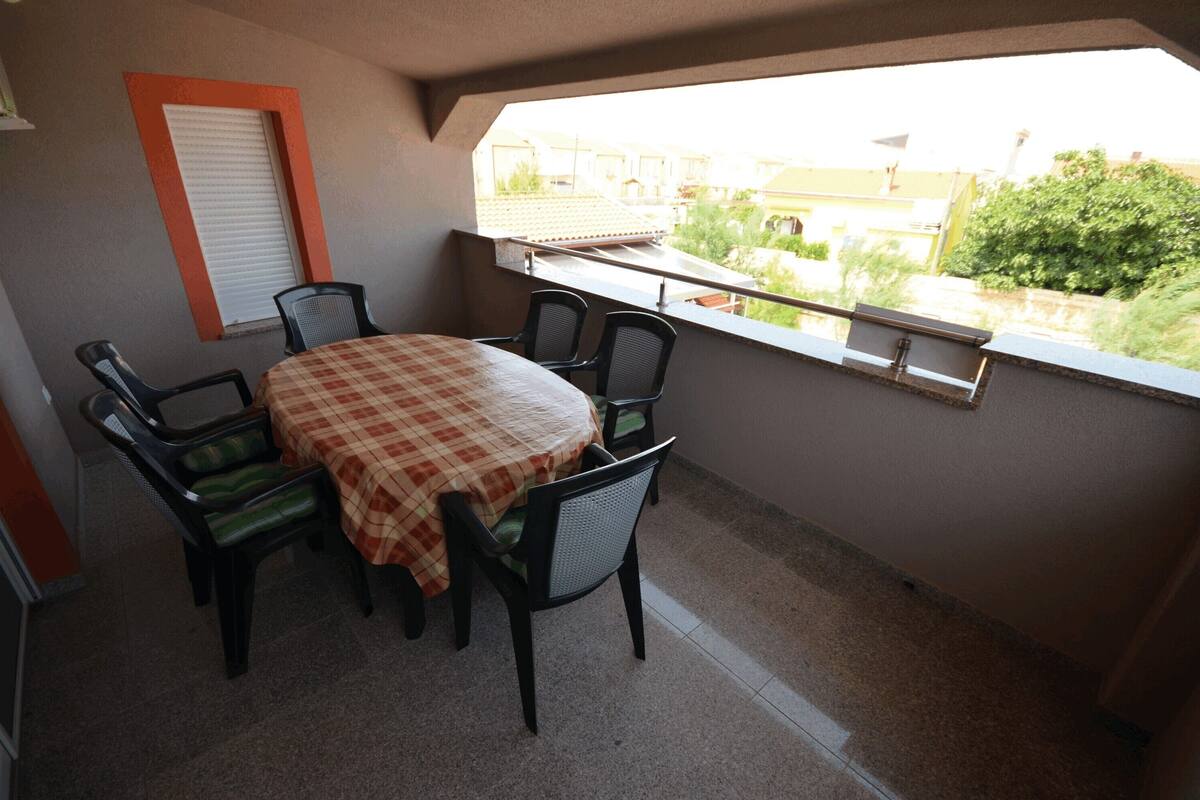 A balcony with a checkered table and chairs, a window with a view of buildings and greenery, and a closed shutter.