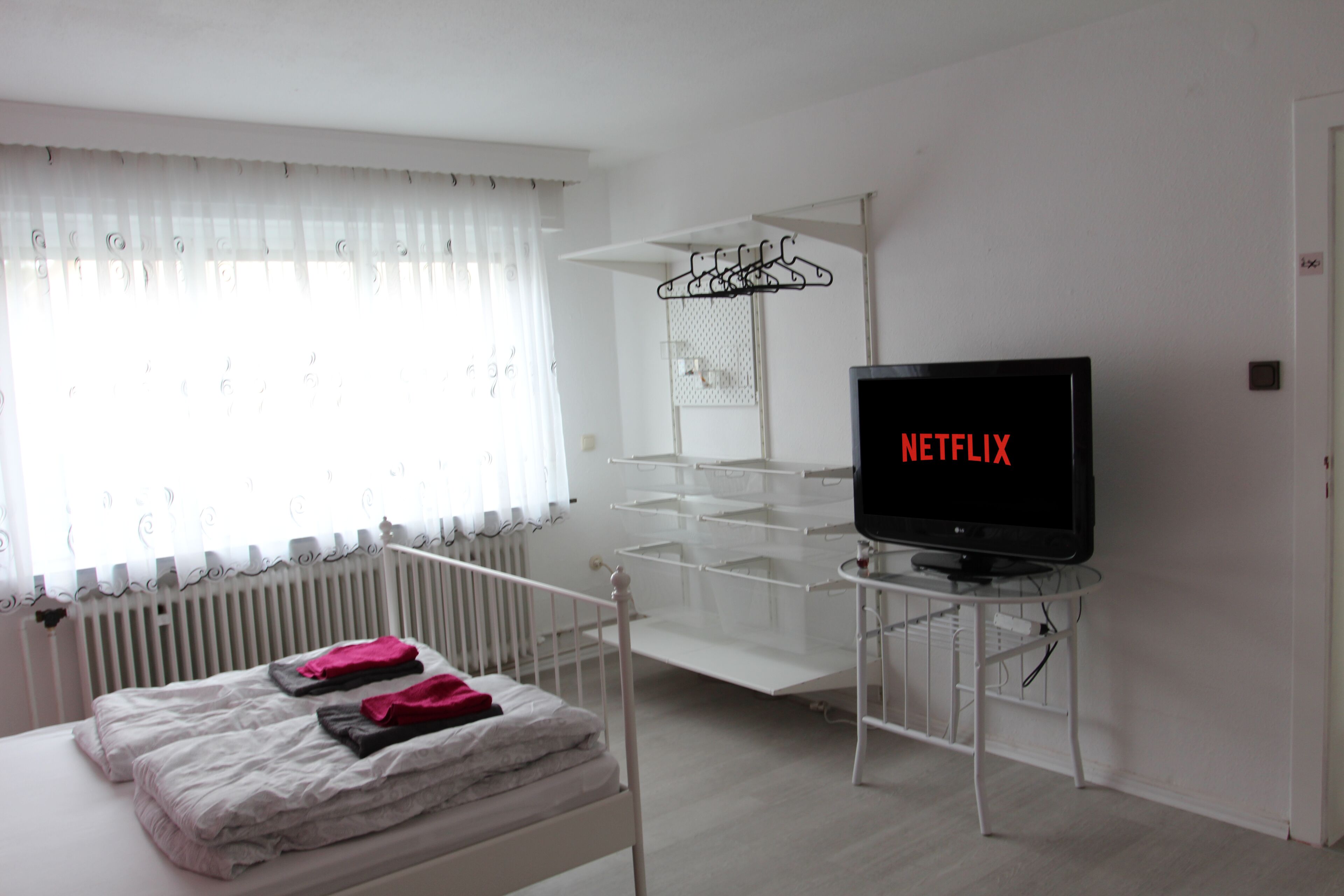 A hotel room with a bed, a TV displaying 'NETFLIX', a radiator, and a window with curtains.