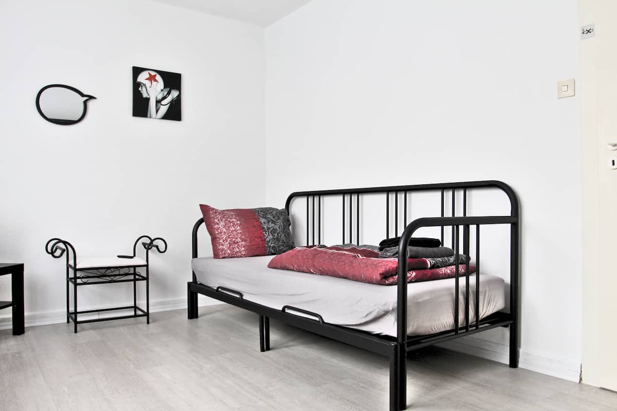 Ett minimalistiskt rum med en svart metallram till sängen, ett litet bord och en stol. Det finns två konstverk som hänger på väggen och en dörr med en ljusströmbrytare.