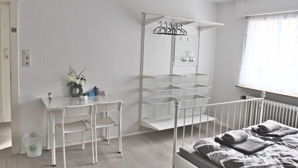 A room with a bed, a desk with chairs, a hanging clothes rack, and a radiator. - Pixie Home (Hanau)