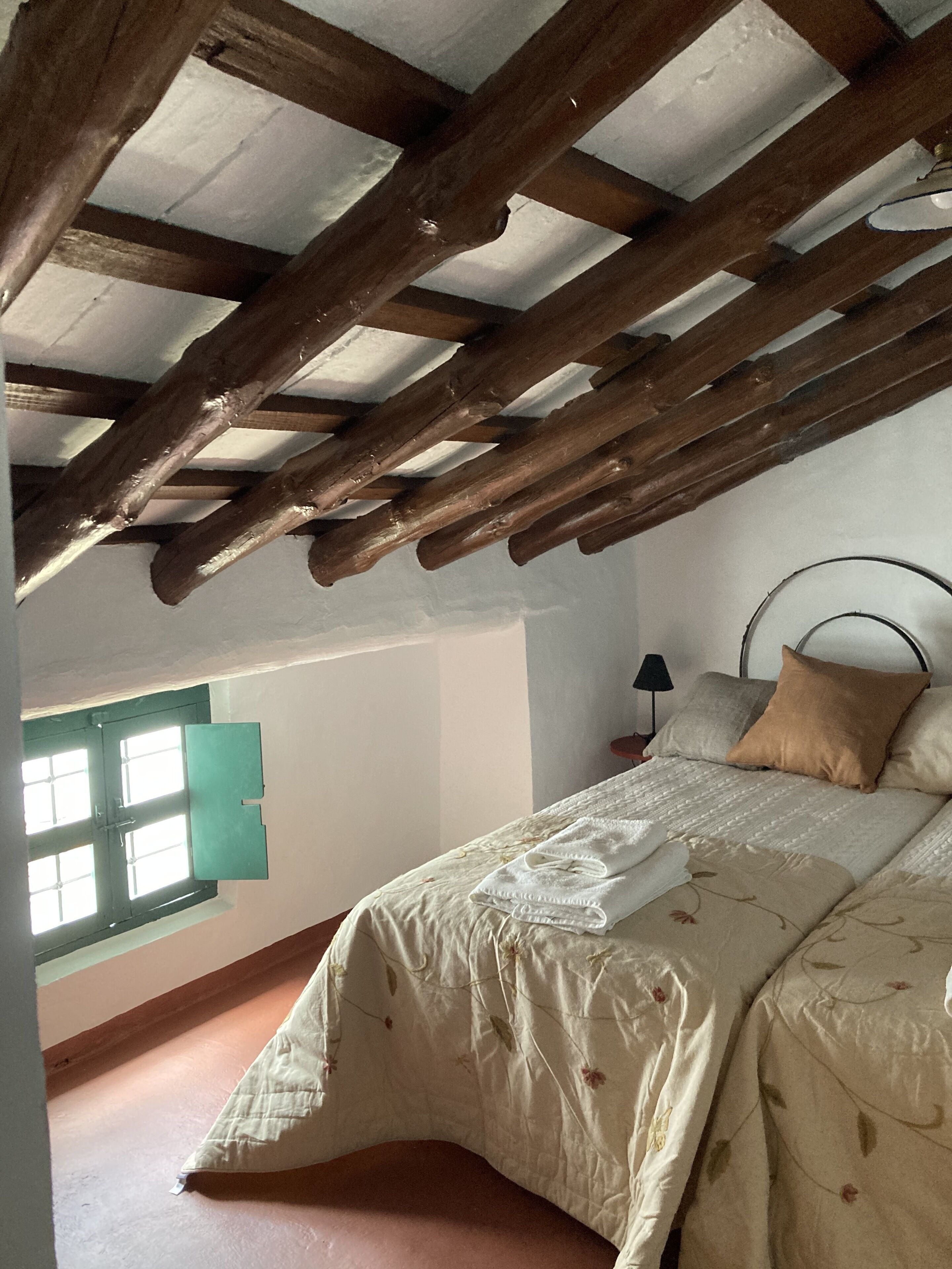 A bedroom with a sloped ceiling, a bed with a floral bedspread, a nightstand with a lamp, and a window with a view of the outside.