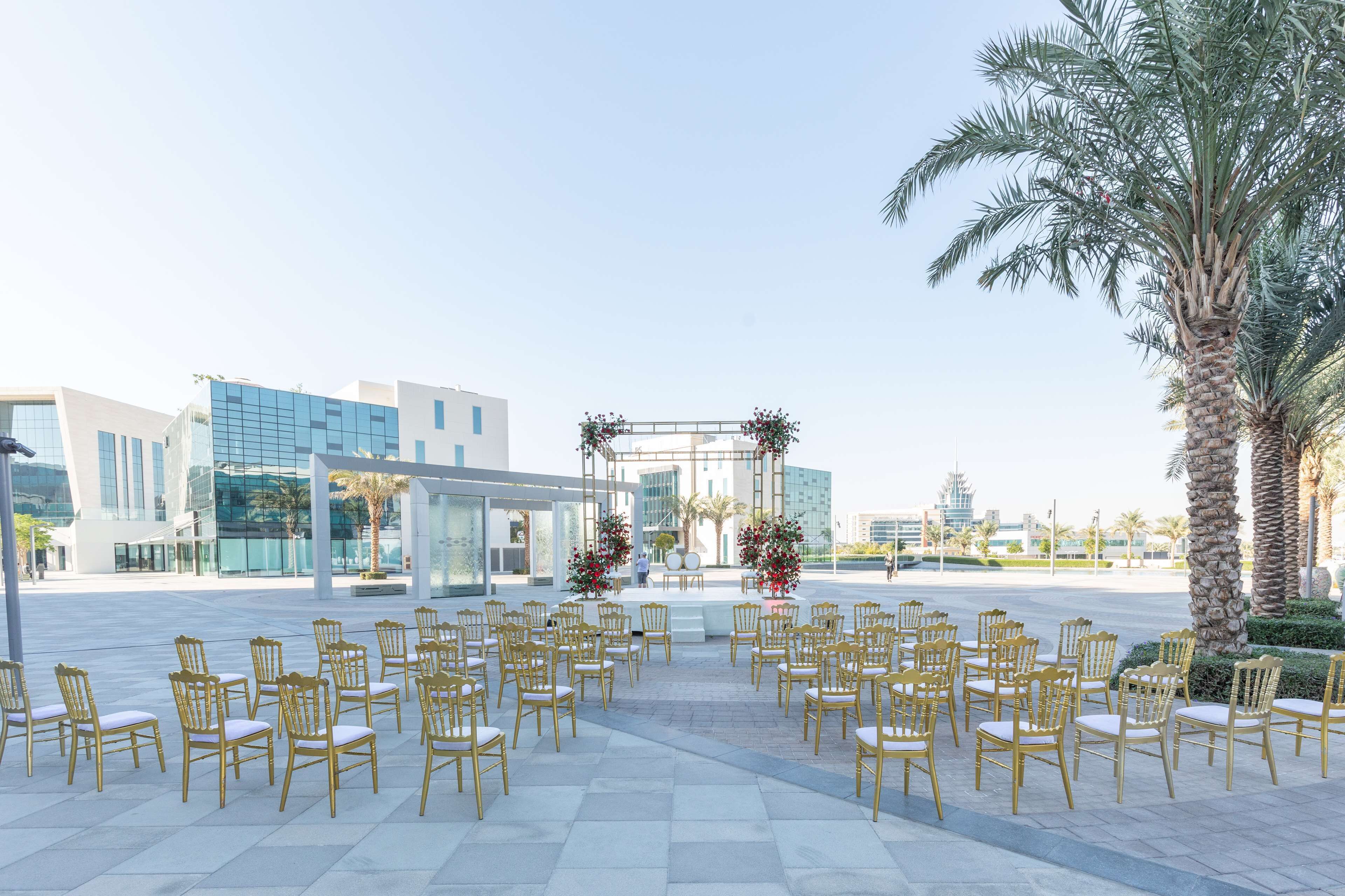 An outdoor seating area with rows of golden chairs arranged for an event, in front of a modern building with glass facades and palm trees.