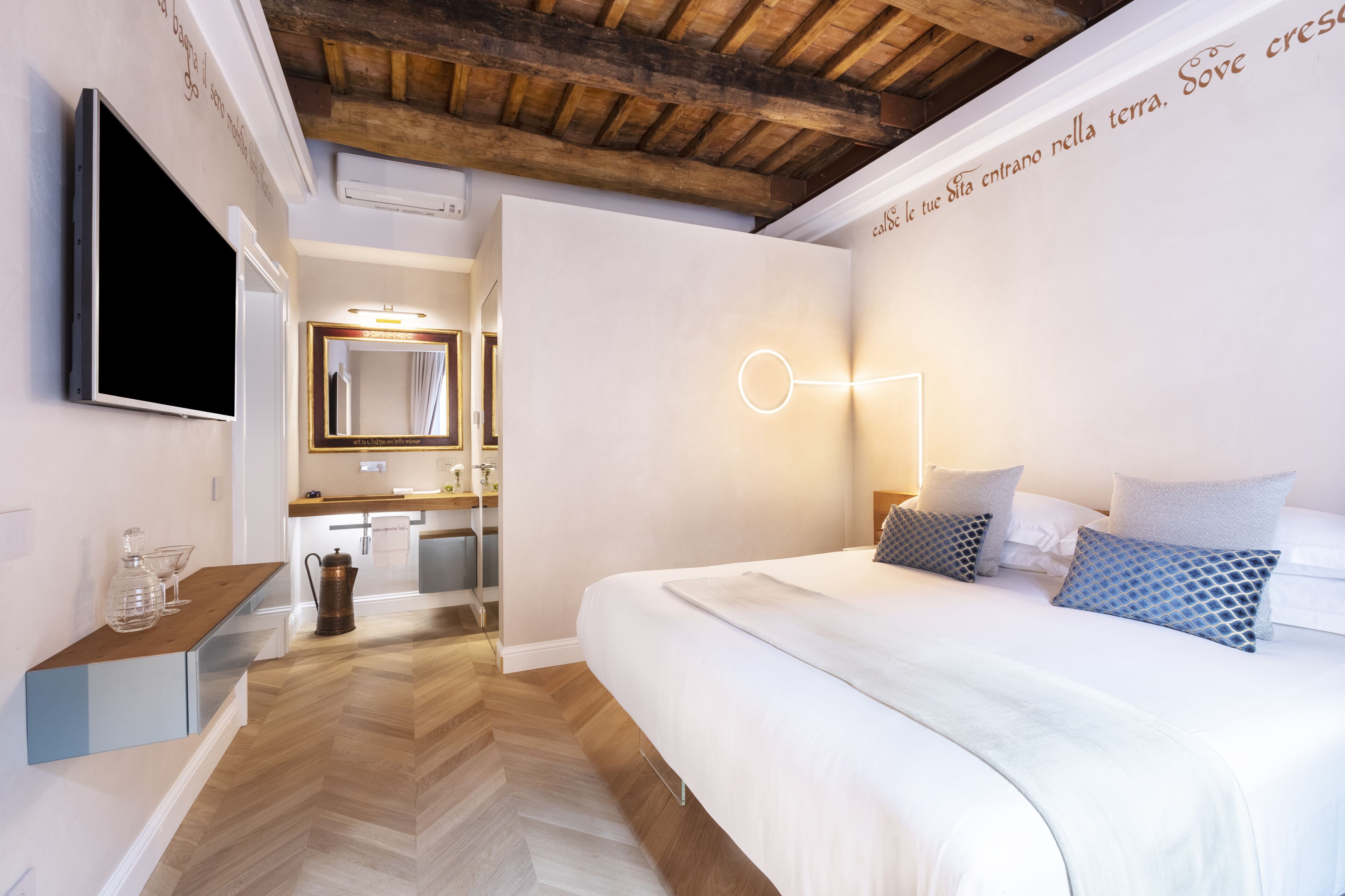 Een moderne hotelkamer met een houten plafond, een groot bed met opvallende kussens, een nachtkastje met een glazen karaf en een flatscreen televisie aan de muur.