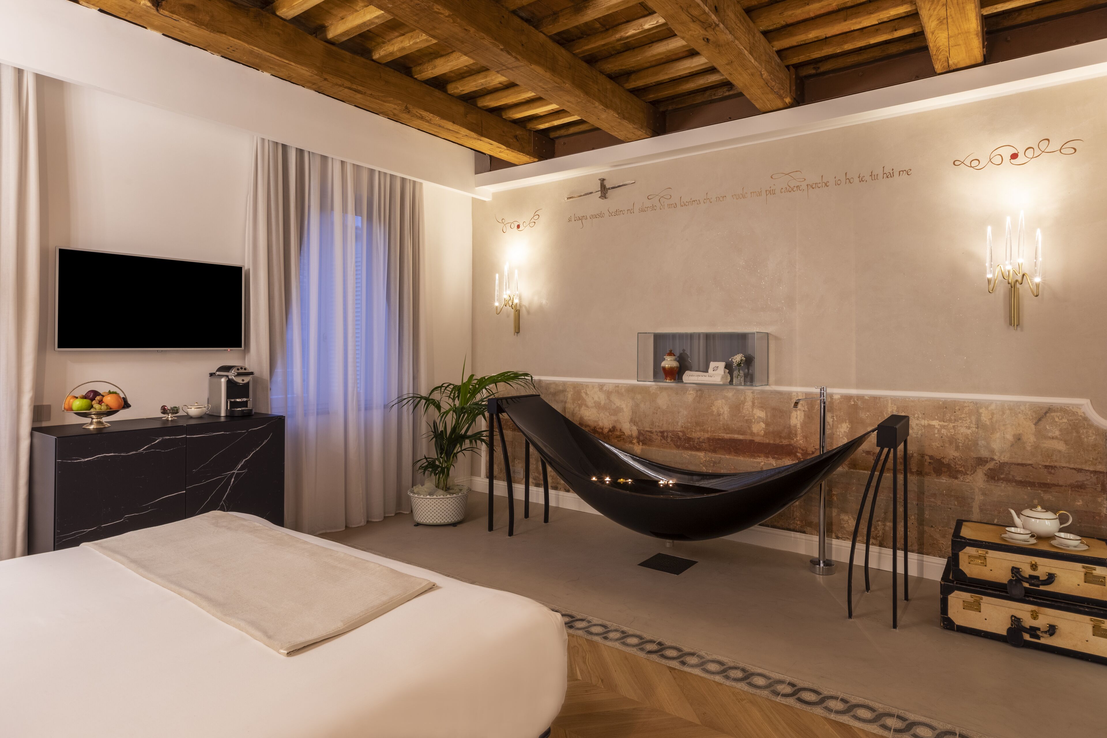 A modern hotel room with a bed, a desk, a black chair, a potted plant, and a wall-mounted TV.
