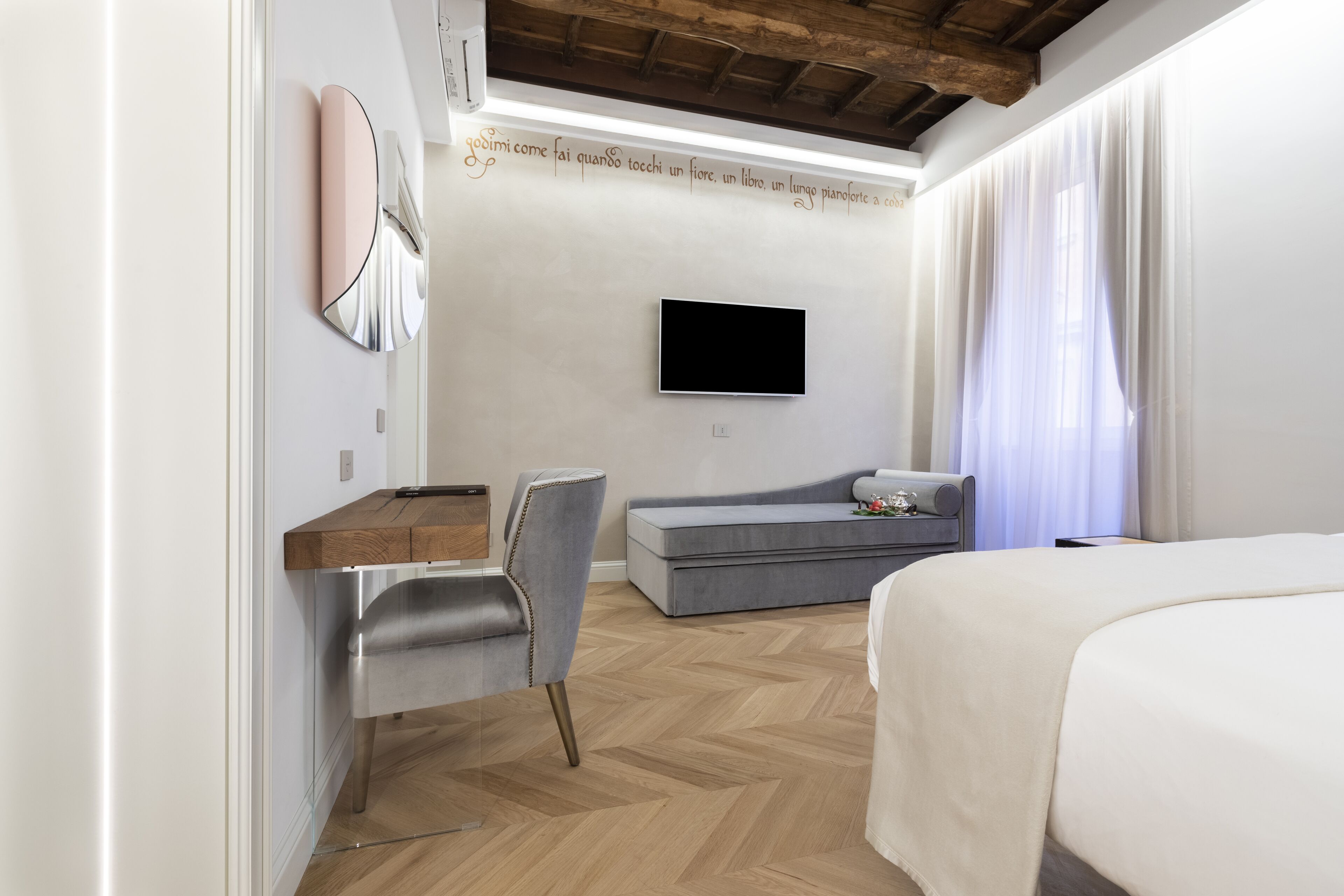 A modern hotel room with a wooden ceiling, a flat-screen TV mounted on the wall, a grey sofa, a wooden desk, and a bed with a white duvet.