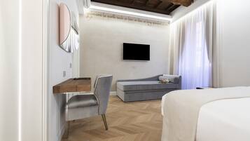 A modern hotel room with a wooden ceiling, a flat-screen TV mounted on the wall, a grey sofa, a wooden desk, and a bed with a white duvet.
