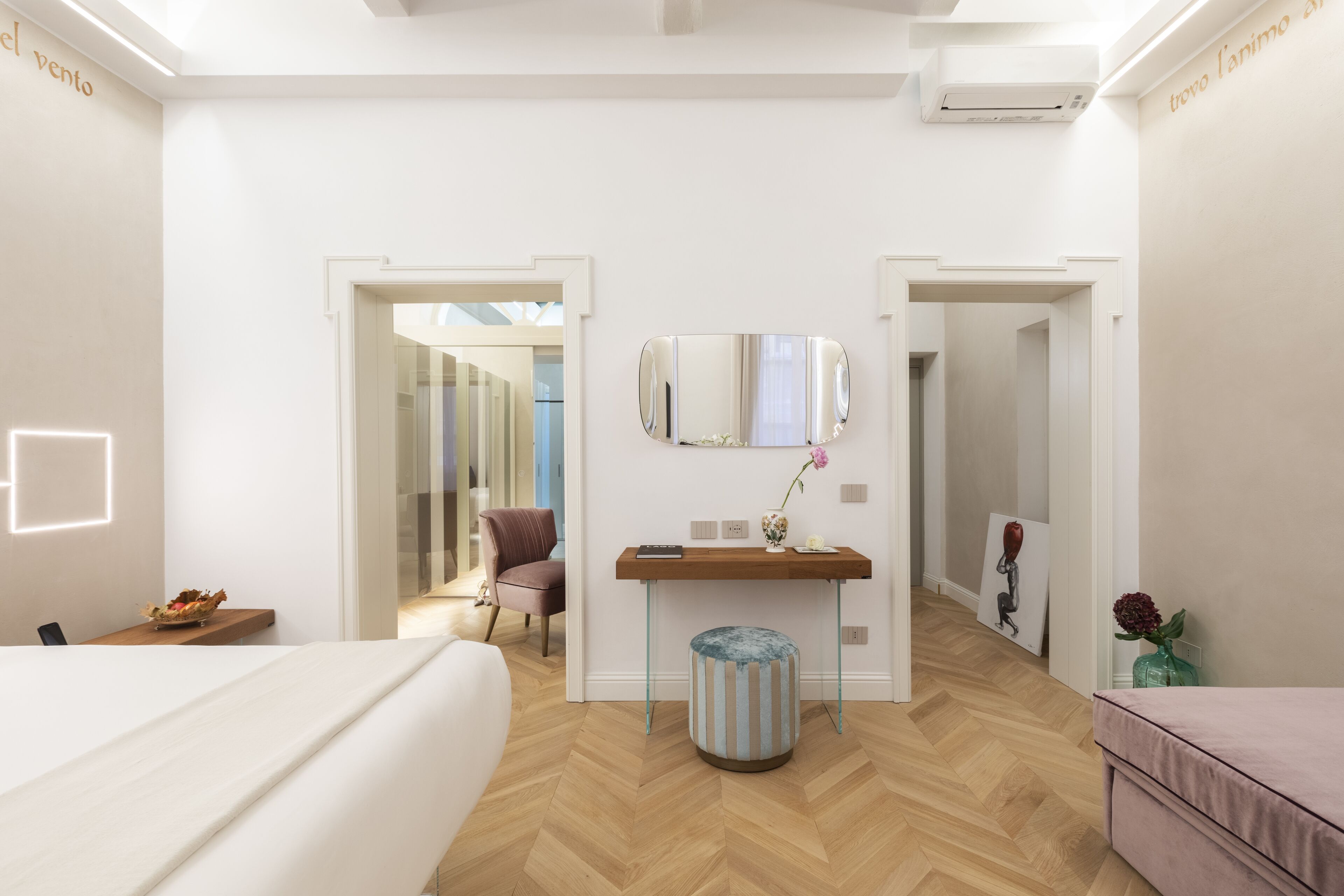 Ein modernes Hotelzimmer mit Holzboden, einem Bett, einem Schreibtisch mit Spiegel, einem kleinen Hocker und einem Gemälde an der Wand.
