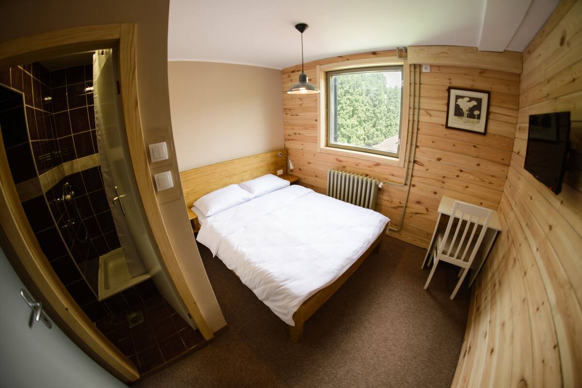 Quarto com cabeceira de madeira, uma cama de solteiro, janela, radiador e televisão na parede.