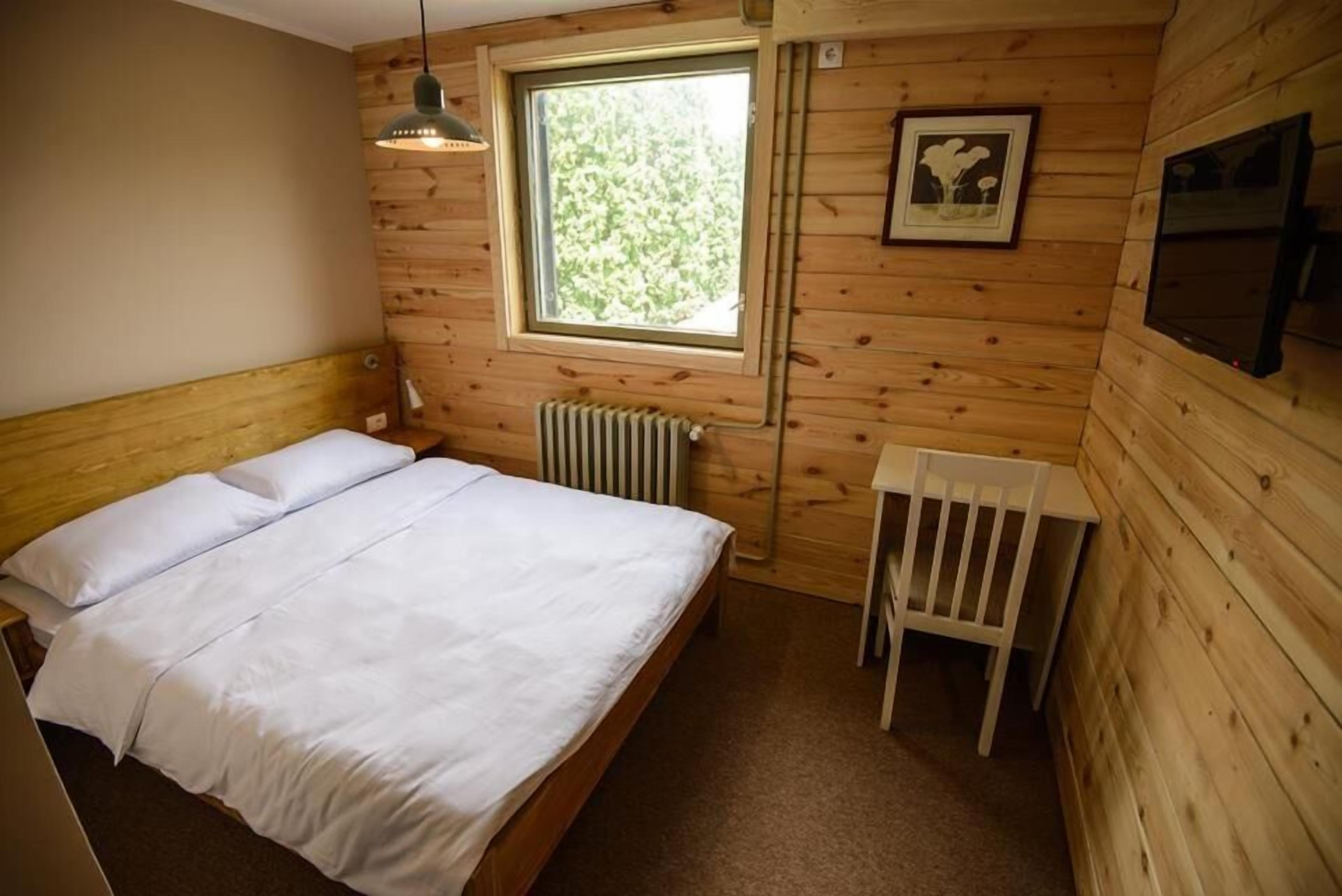A wooden cabin bedroom with a bed, a window, a radiator, and a chair.