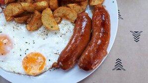 Un plato con salchichas, huevos fritos, cuñas de patata, tomates, rodajas de pepino y un vaso de zumo de naranja.