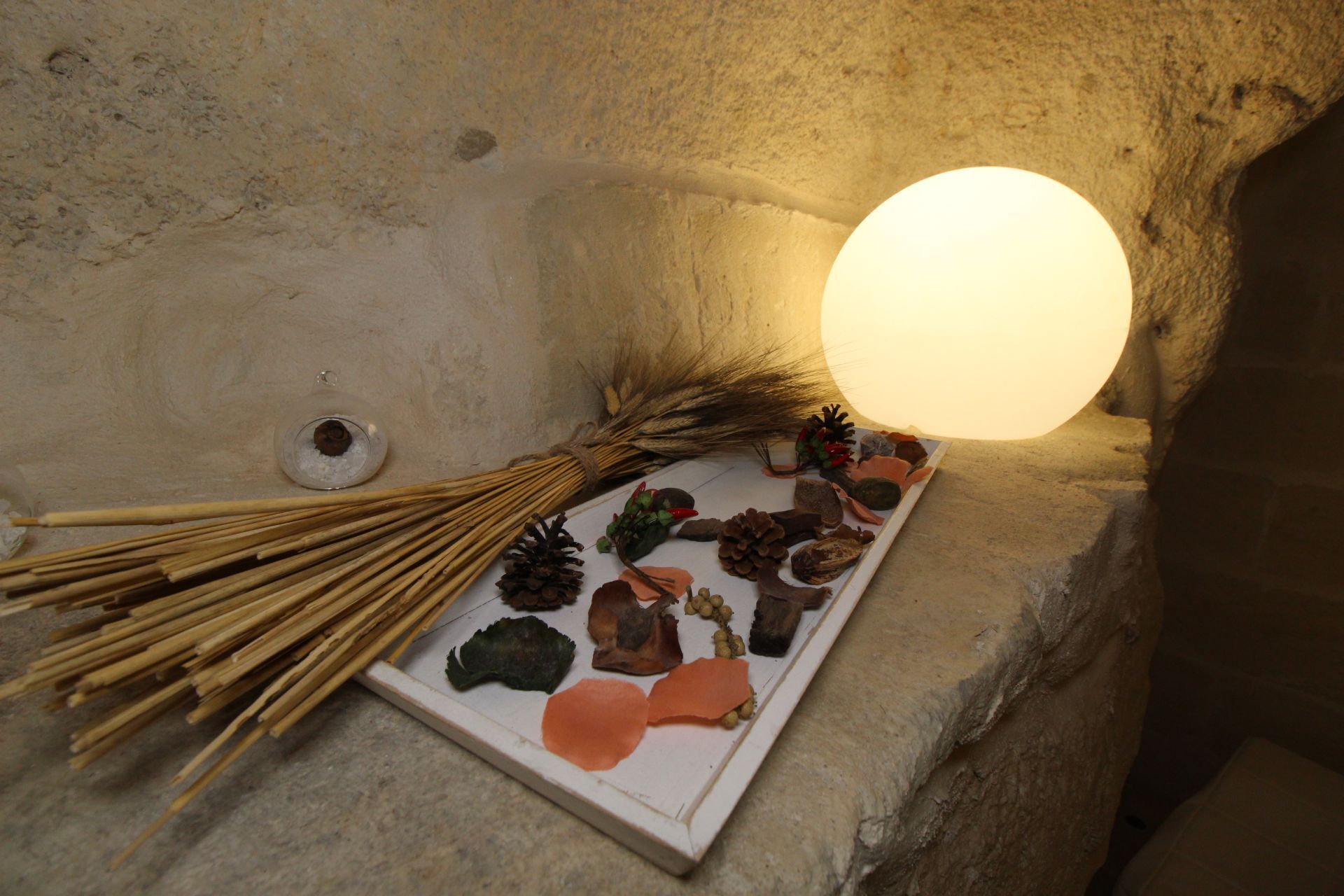 A stone interior with a lit lamp, a small bowl, and a display of natural items like pinecones and leaves.