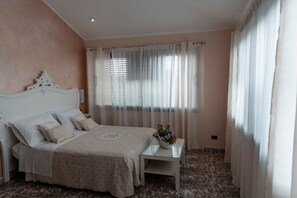Un dormitorio con una cama, una mesita de noche, una pequeña mesa con un florero y cortinas traslúcidas.