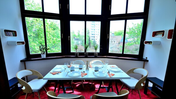 A dining area with a table set for six, surrounded by chairs, large windows with a view of greenery and buildings, and a red carpet. - Victus Apartamenty - Flamingo (Sopot)
