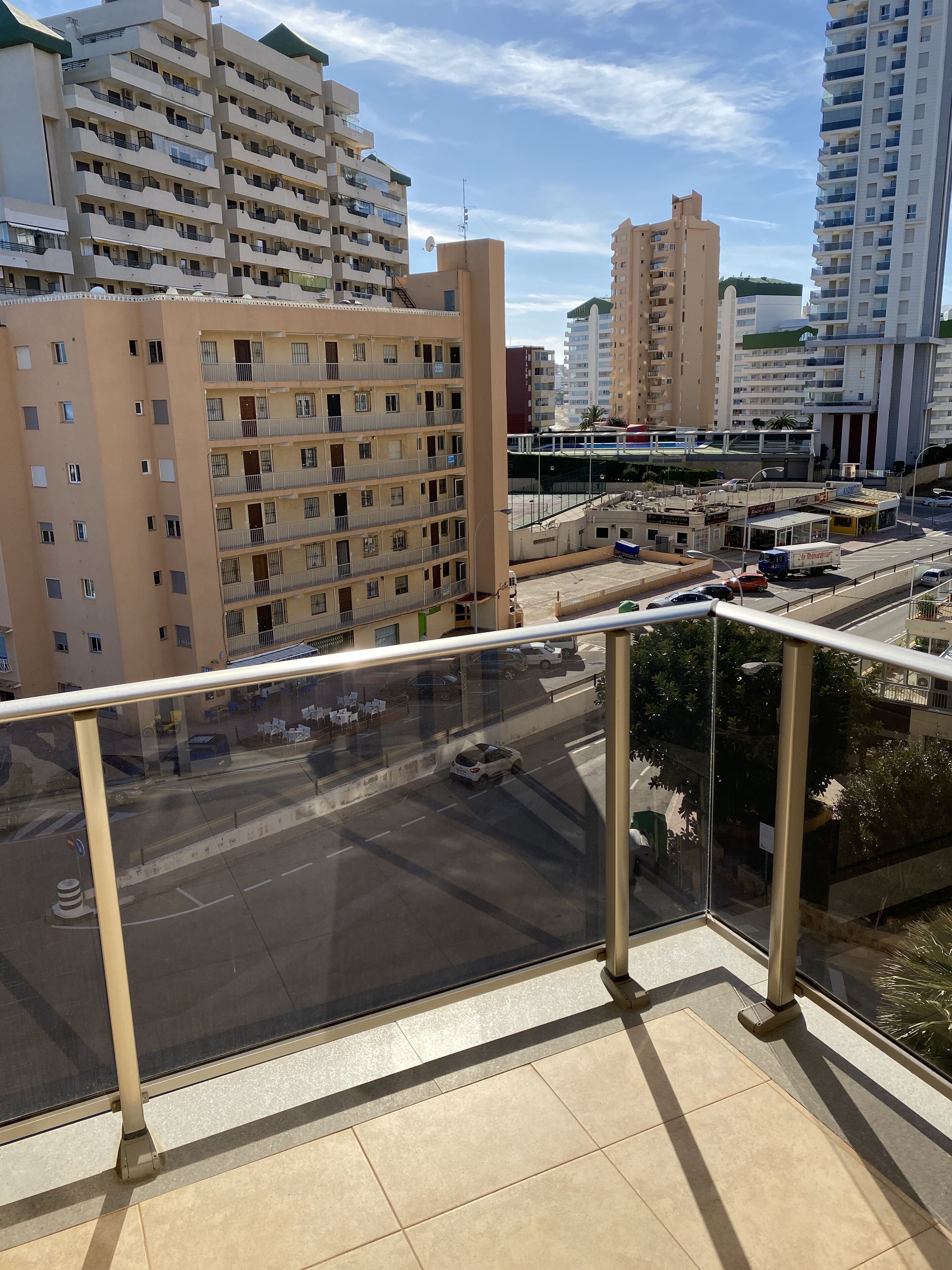A high-rise building with multiple balconies, a parking area with cars, and a clear sky.
