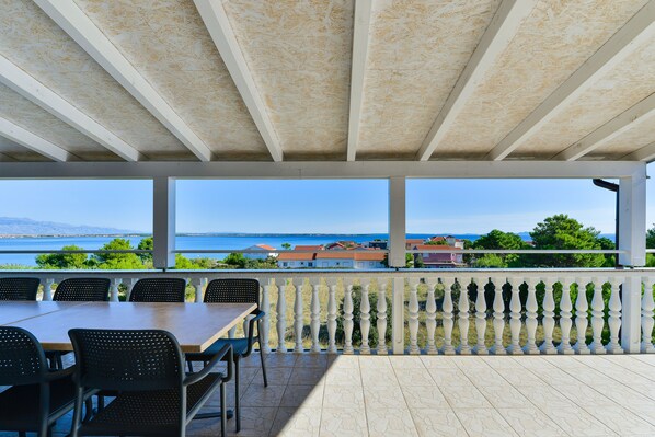 Ein überdachter Außenbereich mit Tisch und Stühlen, mit Blick auf eine malerische Aussicht auf das Meer und Gebäude.