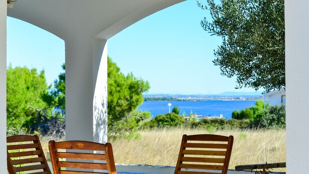 Una mesa y sillas de madera bajo una estructura blanca, con vista al mar y al follaje.