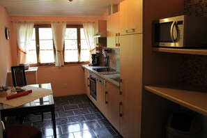 A compact kitchen with a microwave, stove, and sink. There is a dining table with chairs and a window with curtains.