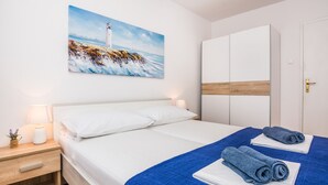 A neatly made bed with white linens and a blue blanket, a bedside table with a lamp, a painting of a lighthouse on the wall, and a wardrobe with a wooden accent panel. - Apartment Poslon (Vir)