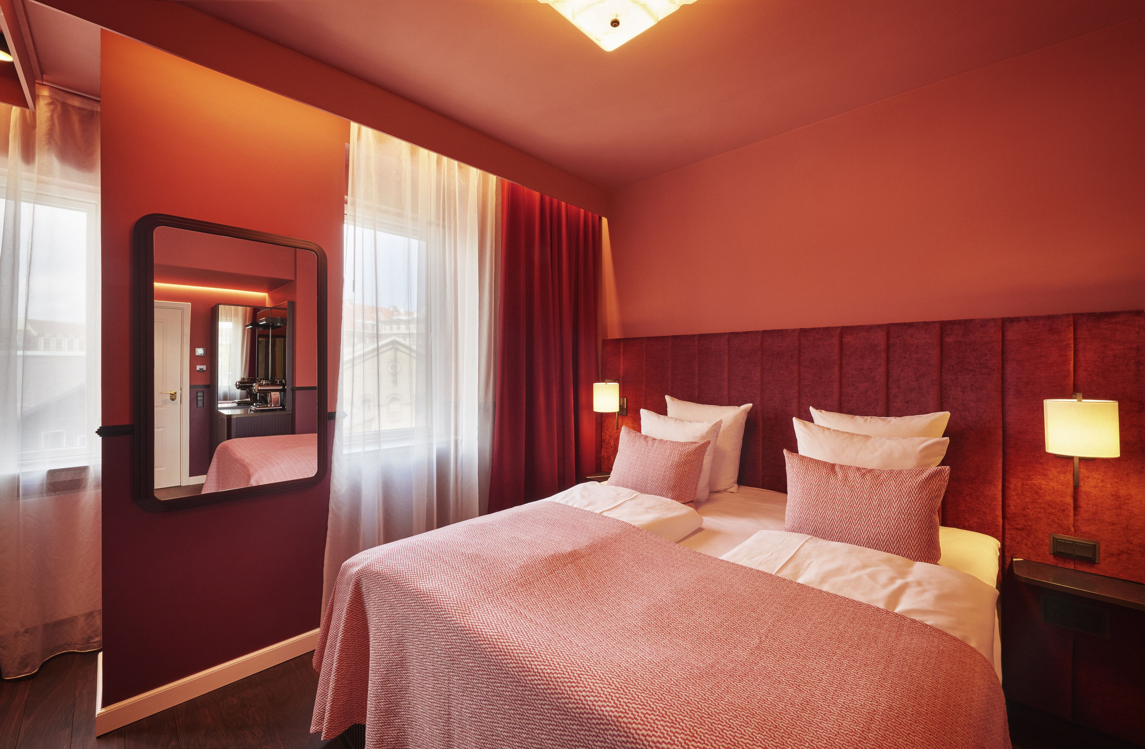 Habitación de hotel con una cama grande, dos mesitas de noche con lámparas, un espejo y vistas a un paisaje urbano a través de cortinas traslúcidas.