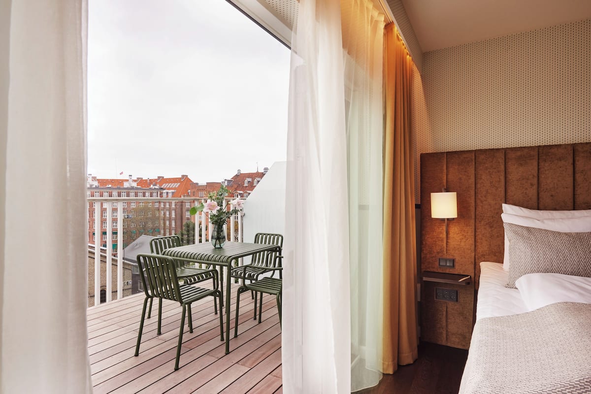 Una habitación de hotel con balcón, cama, mesita de noche y vistas a edificios.