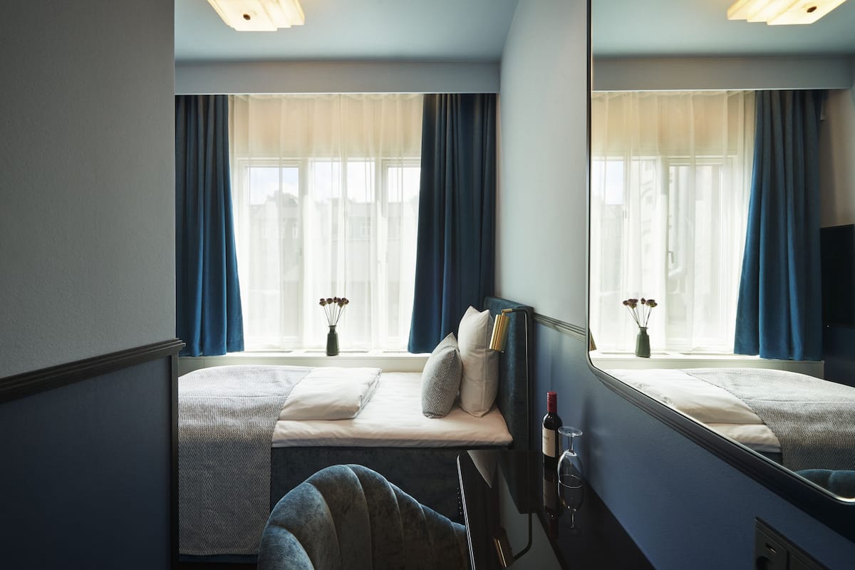 Una habitación de hotel con una cama, una ventana con cortinas, una mesita de noche con una botella de vino y un espejo.