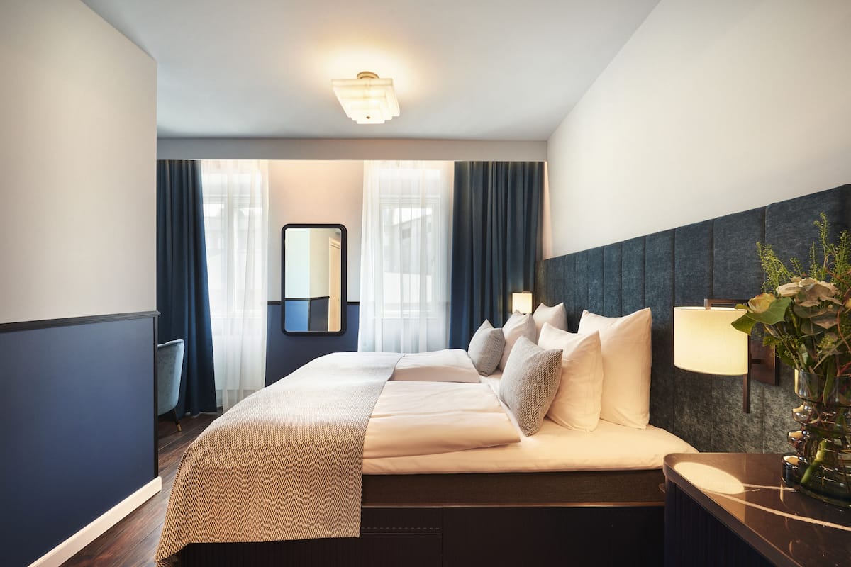 Habitación de hotel con una cama grande, una mesita de noche, un espejo y una ventana con cortinas.