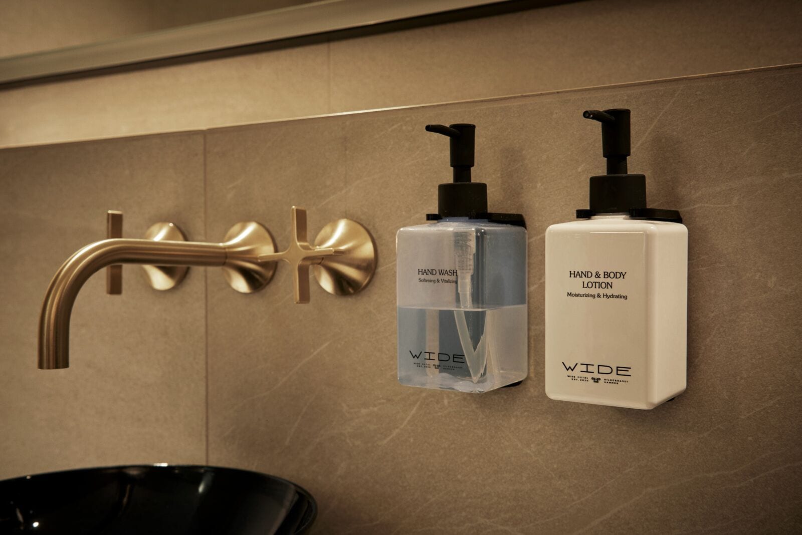 Un baño con un grifo dorado, una botella transparente de jabón líquido y una botella blanca de crema corporal y manos.