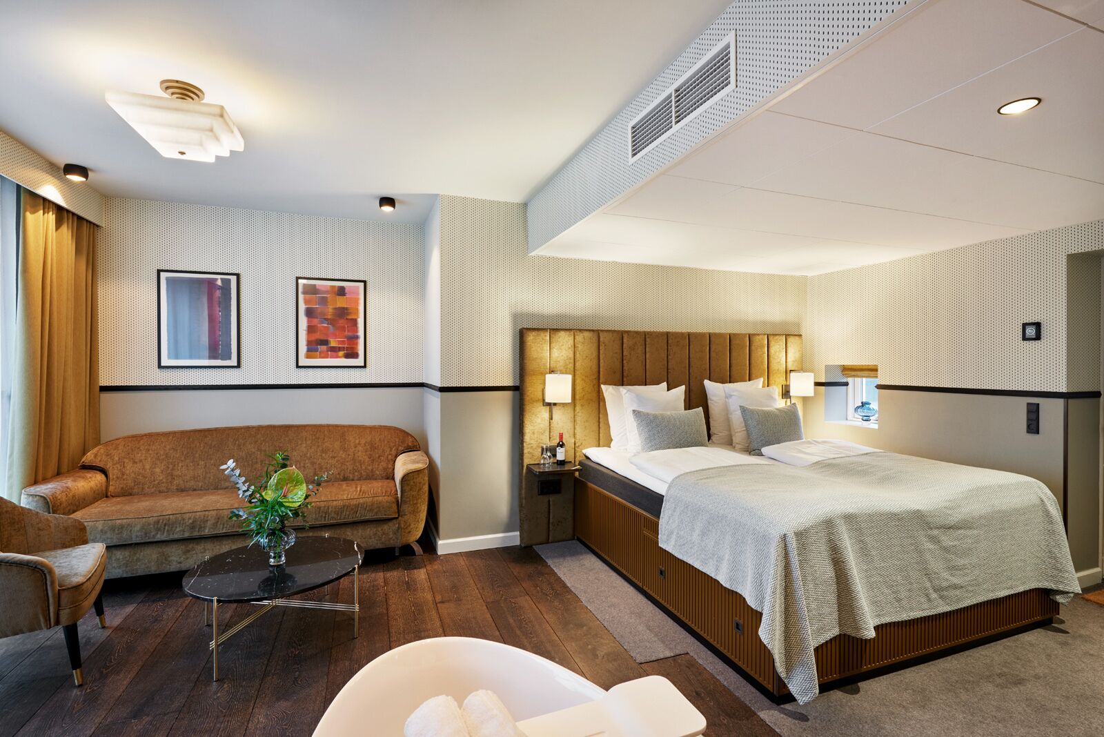 Habitación de hotel con sofá, cama, mesita de noche, lámpara y una pequeña mesa con una planta.