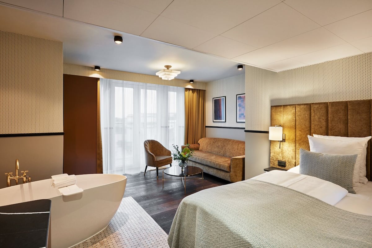 Una habitación de hotel moderna con una cama grande, una zona de estar con sofá y sillón, una mesita con un florero, una bañera y un ventanal con cortinas.